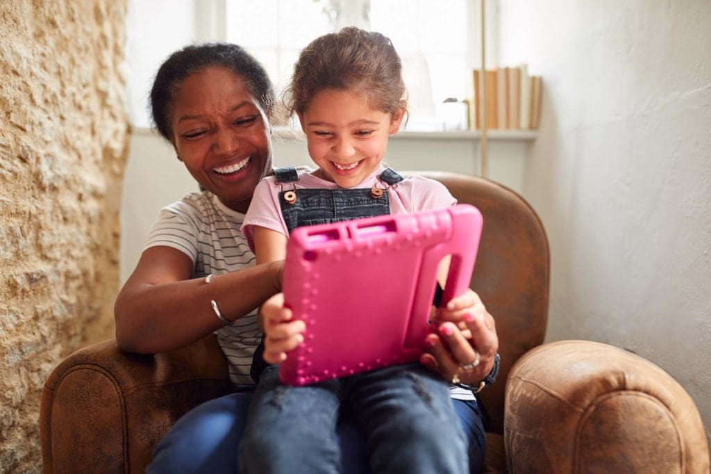Grandmother Sitting In Chair With Granddaughter Watching Movie On Digital Tablet Together
